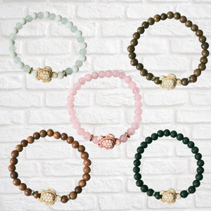 May include: Five beaded bracelets in various colors: light blue, brown, pink, dark brown, and black. Each bracelet features a turtle charm and is displayed against a white brick background. The beads appear to be approximately 0.25 inches in diameter.