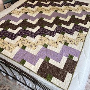May include: A quilted blanket featuring a zigzag pattern composed of fabric blocks in shades of lavender, dark brown, cream, and olive green. The quilt displays a mix of solid colours and floral prints with intricate machine-stitched quilting details.