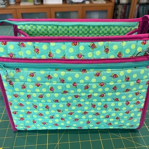 May include: A quilted zippered tote bag with a pink trim and a green and white polka dot pattern. The bag has a small zippered pocket on the front and a handle on the top.