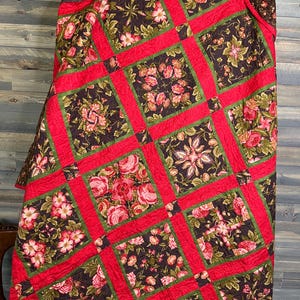Puede incluir: Una colcha acolchada con un estampado floral. La colcha presenta un diseño de patchwork con cuadrados de tela marrón oscuro adornados con flores rosas y blancas. Los cuadrados están enmarcados por bordes rojos y detalles verdes.