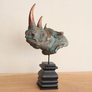 Könnte beinhalten: Eine bronzefarbene Nashornkopfskulptur mit kupferfarbenen Hörnern und einer türkisfarbenen Patina. Der Nashornkopf ist auf einem schwarzen, mehrstufigen Holzsockel montiert, ein dekoratives Element.
