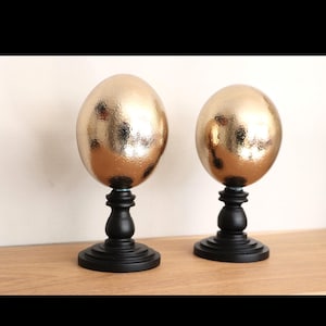 May include: Two decorative golden egg-shaped objects with textured surfaces. Each sits atop a black, ornate pedestal base. The objects are displayed on a light-colored wooden surface, creating a contrast with the gold and black.