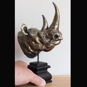 Könnte beinhalten: Bronzefarbene Nashornkopf-Skulptur auf einem schwarzen Holzsockel. Das Nashorn hat detaillierte Texturen und markante Hörner. Die Skulptur wird auf einem Ständer präsentiert.