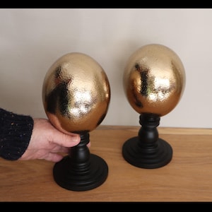 May include: Two decorative golden egg sculptures with textured surfaces, mounted on black wooden pedestals. The eggs have a metallic finish, reflecting light. The pedestals have a classic, turned design, adding a touch of elegance to the home decor.