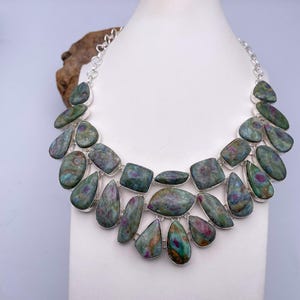 May include: A multi-layered statement necklace featuring polished, teardrop and rectangular-shaped green and magenta zoisite stones set in silver-toned metal bezels. The stones are arranged in two rows, connected by a silver-toned chain link necklace.