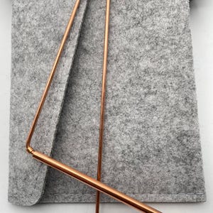 May include: Copper-colored metal dowsing rods, with a triangular shape, rest on a gray felt pouch. The rods are thin and shiny, with a slight bend at the ends. The pouch has a black button closure.