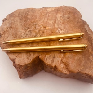 May include: Two gold-colored metal ballpoint pens with a smooth, cylindrical barrel, a pointed tip, and a slim clip attached to the top. The pens feature a polished finish and are positioned horizontally, one above the other, with a uniform design.