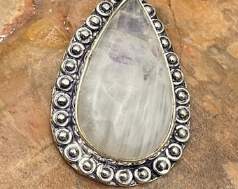 May include: A teardrop-shaped pendant with a large, pearly white stone. The stone has subtle internal striations and a hint of purple. The pendant is framed by a silver-colored metal with a beaded border and a decorative bail.