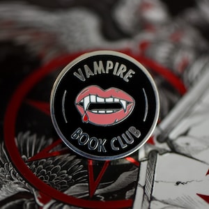 May include: A black and silver enamel pin with a red vampire mouth and fangs. The pin says "Vampire Book Club" in white letters.