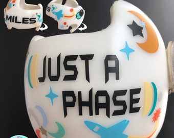 May include: A white, space-themed baby bottle with the text "JUST A PHASE" in black. The bottle is decorated with a blue rocket, stars, and a crescent moon. A small, matching pacifier is also visible, with the name "MILES" on it.