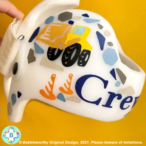 May include: White helmet with a yellow construction truck, orange trees, and blue and grey geometric shapes. The name "Crew" is written in blue letters on the helmet.