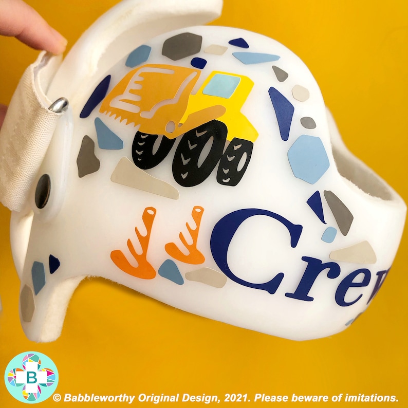 May include: White helmet with a yellow construction truck, orange trees, and blue and grey geometric shapes. The name "Crew" is written in blue letters on the helmet.
