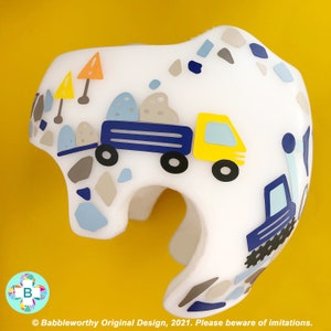 May include: A white helmet featuring a blue and yellow truck carrying rocks, a blue truck with a caterpillar track, and a pattern of grey and blue stones. The helmet is decorated with a construction theme.