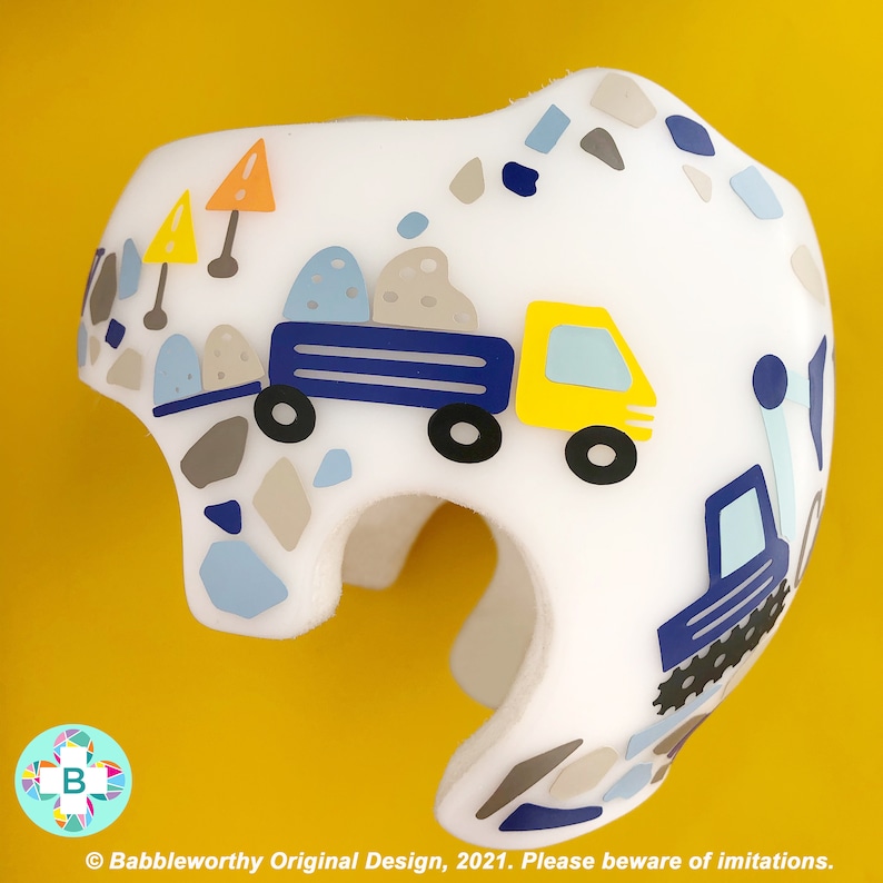 May include: A white helmet featuring a blue and yellow truck carrying rocks, a blue truck with a caterpillar track, and a pattern of grey and blue stones. The helmet is decorated with a construction theme.