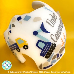 May include: White helmet with a blue, yellow and grey construction themed design. The helmet has the words "Under Construction" written on it.