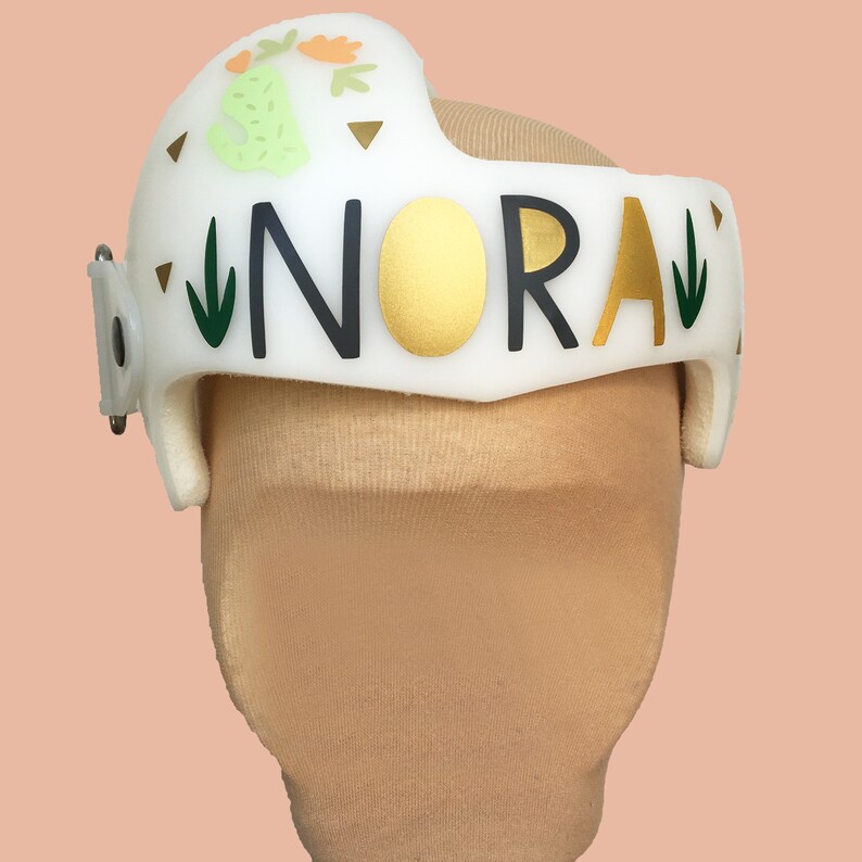 Puede incluir: Casco blanco con un dise&ntilde;o 'NORAH' en oro y negro, acentos de cactus verde y flores, y una correa blanca.