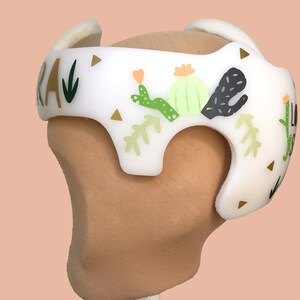 Puede incluir: Casco acolchado blanco con un dise&ntilde;o de cactus verde, negro y dorado. El casco tiene la palabra "RA" escrita en el lateral.