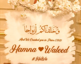 May include: A white acrylic sign with a gold brushstroke design. The sign features Arabic calligraphy, the text "And We Created you in Pairs (788)", and the names "Hanna" and "Waleed" with a heart symbol between them. The hashtag "#HarWa" is also included. The sign is displayed on a wooden easel with white flowers.
