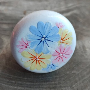 May include: A white, round decorative knob with hand-painted floral designs. The flowers are in shades of blue, pink, and yellow, with orange centers. The knob is likely made of wood and is suitable for furniture or cabinet hardware.