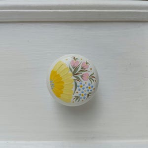 Könnte beinhalten: Ein weißer runder Knopf mit handbemaltem Blumendesign. Das Design zeigt gelbe, rosa und blaue Blumen.