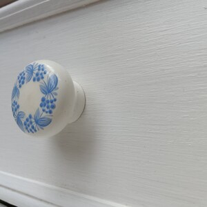 May include: White ceramic drawer knob with a blue floral design. The knob has a raised edge and a smooth surface.