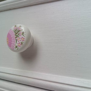 May include: White ceramic drawer knob with a pink floral design. The knob has a round shape and a smooth surface.