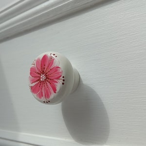 Op de afbeelding: Witte ladeknop met een handgeschilderd roze bloemmotief. De bloem heeft een gedetailleerd midden en roze bloemblaadjes. De knop is bevestigd op een witte lade.