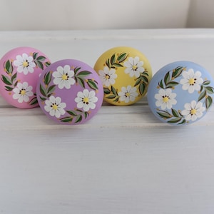 May include: Four round knobs with floral designs. The knobs are painted in shades of pink, purple, yellow, and blue. Each knob features a wreath of white flowers with green leaves.