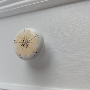 May include: A round, white drawer knob with a painted yellow flower design. The flower has a brown center and brown lines radiating out from the center.