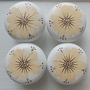 May include: Four white wooden knobs with a painted yellow flower design. Each knob has a brown centre with white dots and brown stems with small white dots.