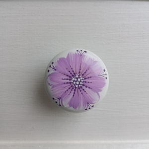 May include: A white ceramic drawer pull with a hand-painted purple flower design. The flower has a detailed center and delicate petal strokes. The drawer pull is round and sits on a white surface.