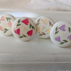 Op de afbeelding: Vier keramische knoppen met bloemmotieven. De knoppen zijn wit met roze, gele, paarse en bruine bloemen die erop geschilderd zijn.