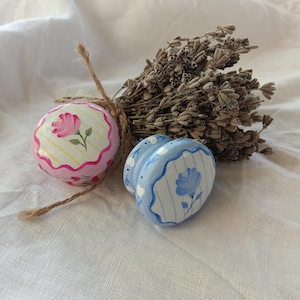 Op de afbeelding: Een paar decoratieve keramische zout- en peperstrooiers, één roze en één blauw, met bloemmotieven. De roze strooier heeft een roos, terwijl de blauwe strooier een blauwe bloem heeft. Een bosje gedroogde lavendel is vastgebonden met touw.