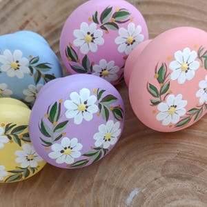 以下が含まれることがあります： 花柄が手描きされた木製ノブが4つ。ノブはパステルピンク、ブルー、イエロー、パープルでペイントされています。各ノブには、黄色の芯を持つ白いデイジーが、緑の葉に囲まれています。