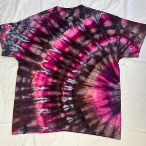 May include: A short-sleeve t-shirt featuring a vibrant tie-dye pattern in shades of hot pink, deep brown, and black. The design radiates from the lower right side in a circular fan motif. A small white tag on the side clearly displays the text XL.
