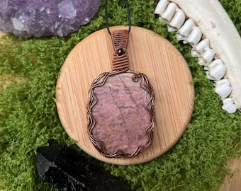 May include: A handcrafted pendant featuring a pink rhodonite stone wrapped in copper wire. The pendant hangs on a black cord, set against a wooden disc and green moss. A black crystal and amethyst cluster are also visible.