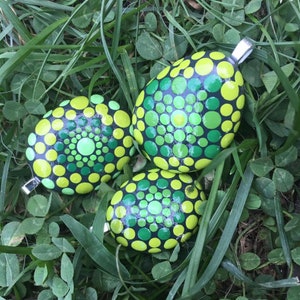 May include: Three hand-painted, oval-shaped pendants with silver-colored bails, resting on grass. Each pendant features a black base with a pattern of green and yellow dots. The dots vary in size, creating a mandala-like design.