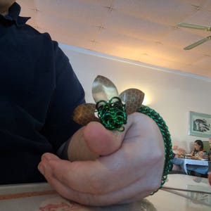 May include: A green and gold chainmail bracelet with a flower design. The bracelet is made of metal rings and has a textured finish.
