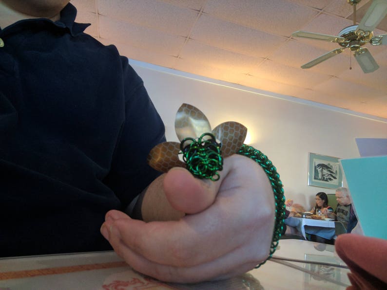 May include: A green and gold chainmail bracelet with a flower design. The bracelet is made of metal rings and has a textured finish.