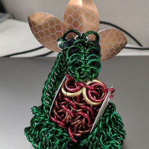 May include: A green, red, and gold chainmail sculpture with a copper-colored flower on top. The sculpture is made of small metal rings woven together.