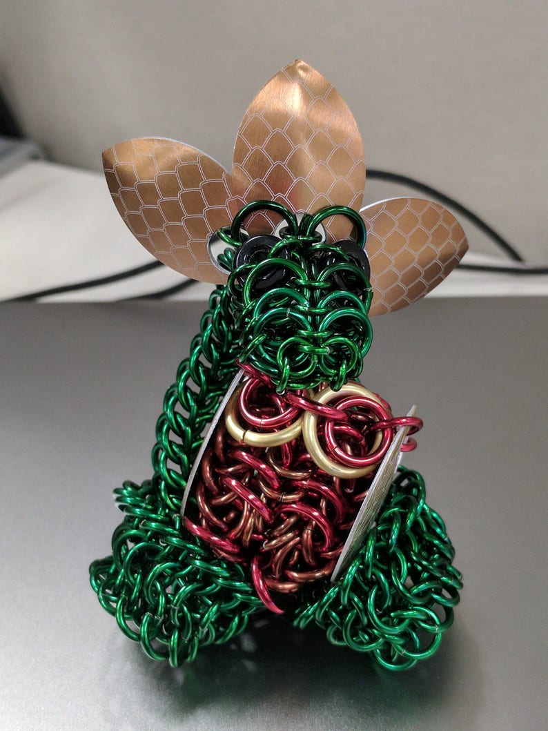 May include: A green, red, and gold chainmail sculpture with a copper-colored flower on top. The sculpture is made of small metal rings woven together.