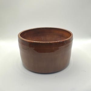 May include: A cylindrical ceramic bowl with a smooth, glossy finish in a deep, warm brown color. The vessel features straight, vertical sides and a wide, open top, showcasing a consistent, reflective glaze across its entire exterior surface.