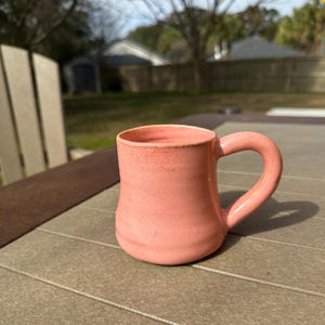 May include: A handmade ceramic mug in a soft coral color. The mug has a rounded body, a curved handle, and a slightly textured surface. It sits on a brown and gray outdoor table, with a blurred background of trees and a house.