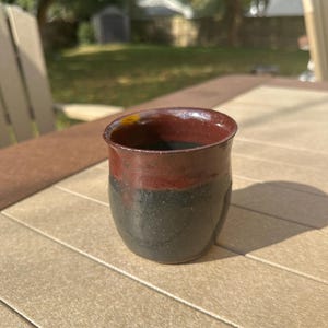 May include: A handmade ceramic tumbler with a dark red rim and a speckled black and dark green body. The cup is sitting on a light brown table in natural sunlight.