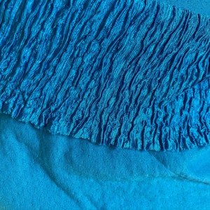 May include: Close-up of a vibrant turquoise fabric. The top layer features a textured, ruffled design, while the bottom layer is a smooth, solid turquoise. The contrasting textures and shades create visual interest. The image highlights the fabric's color and detail.
