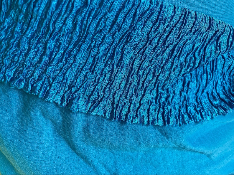 May include: Close-up of a vibrant turquoise fabric. The top layer features a textured, ruffled design, while the bottom layer is a smooth, solid turquoise. The contrasting textures and shades create visual interest. The image highlights the fabric's color and detail.