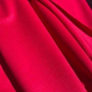 May include: Close-up of vibrant red fabric with soft folds and subtle shadows. The material appears smooth and possibly silky, with a rich, saturated color. The folds create a sense of depth and texture.