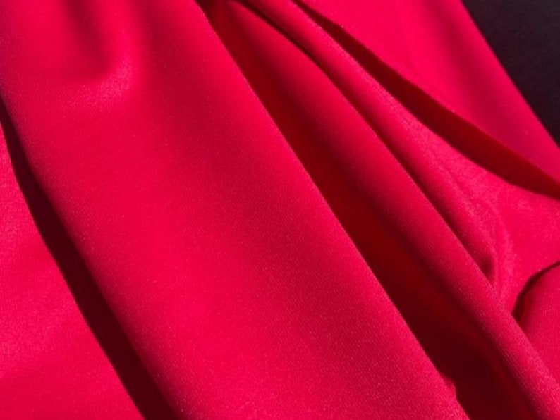 May include: Close-up of vibrant red fabric with soft folds and subtle shadows. The material appears smooth and possibly silky, with a rich, saturated color. The folds create a sense of depth and texture.