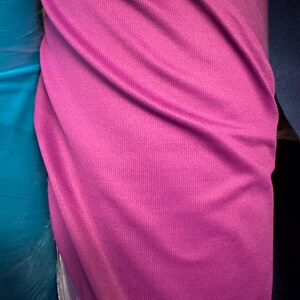 May include: A roll of vibrant magenta fabric, tightly wrapped in clear plastic. The fabric has a ribbed texture, with visible vertical lines. The color is a rich, saturated shade of pink, suitable for various textile projects.