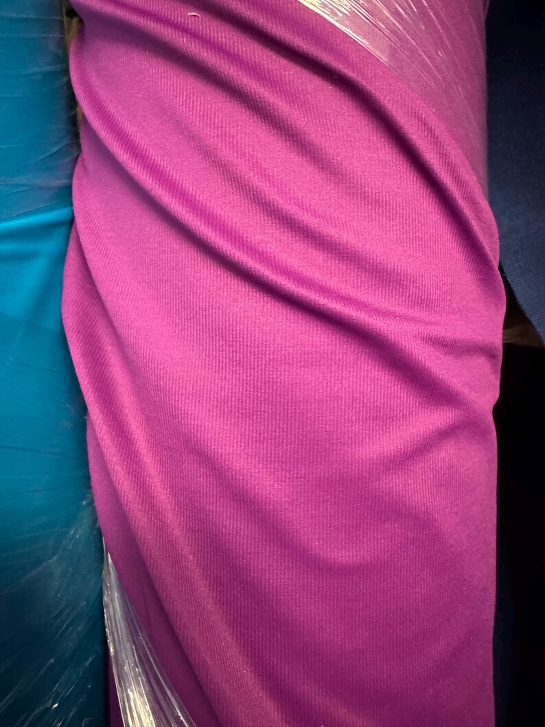 May include: A roll of vibrant magenta fabric, tightly wrapped in clear plastic. The fabric has a ribbed texture, with visible vertical lines. The color is a rich, saturated shade of pink, suitable for various textile projects.